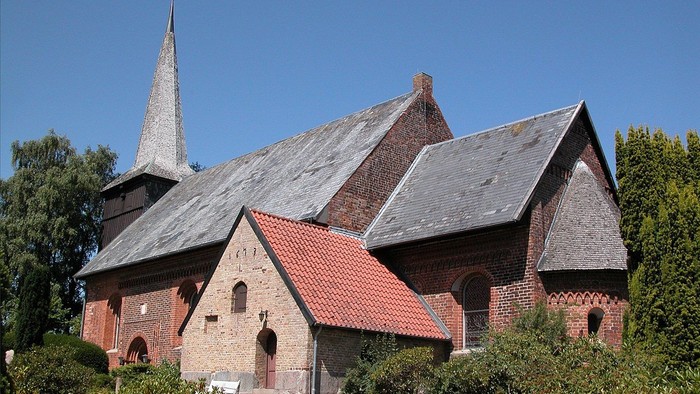 Kirchengebäude mit spitzem Turm und rotem Dach, umgeben von Grün.