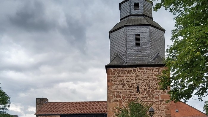 Historische Steinkirche mit modernem Glasanbau, umgeben von Grünflächen und vor bewölktem Himmel gelegen.