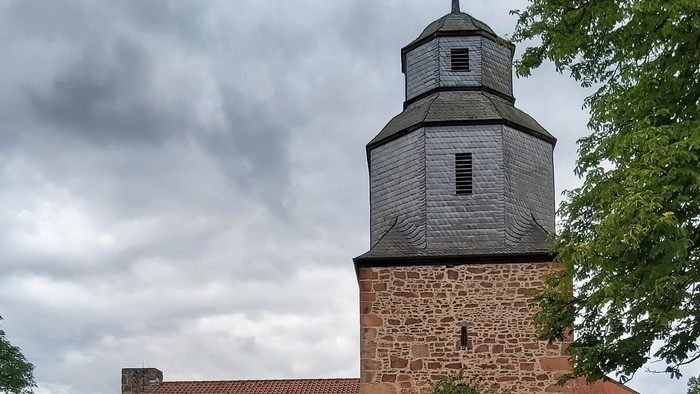 Historische Steinkirche mit modernem Glasanbau, umgeben von Grünflächen und vor bewölktem Himmel gelegen.