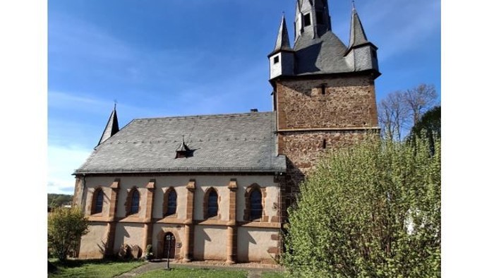 Kirche mit hohem Turmspitz und Rundbogenfenstern, umgeben von grünem Gras und Sträuchern.