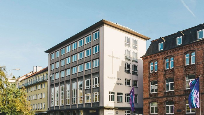 Moderner Bau mit Kreuzsymbol, umgeben von älteren Backsteingebäuden unter klarem Himmel.