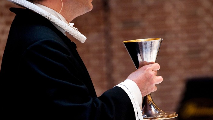 En person i ceremonielt tøj holder en liturgisk kalk udendørs.