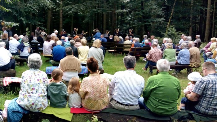 Eine große Menschenansammlung im Freien, die auf Bänken sitzt und einer Aufführung in einem bewaldeten Gebiet lauscht.