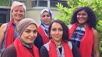 Fünf Frauen mit roten Schals und Kopftüchern posieren gemeinsam im Freien in der Nähe von Grünflächen.