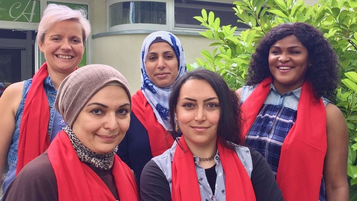 Fünf Frauen mit roten Schals und Kopftüchern posieren gemeinsam im Freien in der Nähe von Grünflächen.