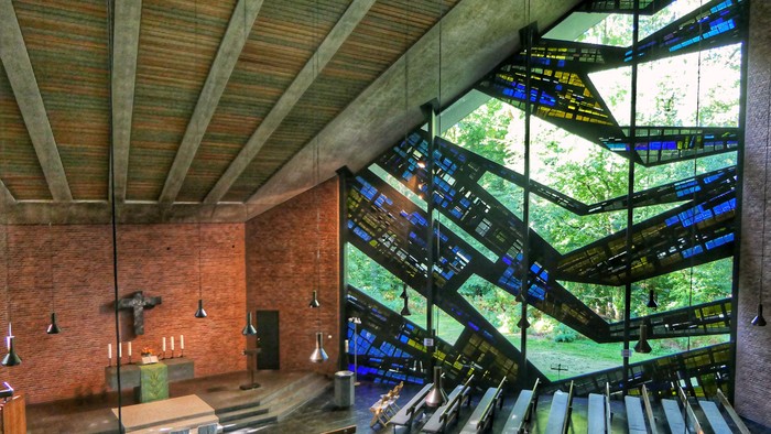 Moderne Kapelle mit geometrischen Buntglasfenstern und Holzbänken unter einer Holzdecke.