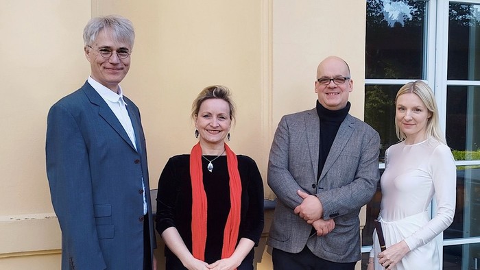 Vier Personen posieren formell vor einem Gebäudeeingang in professioneller Kleidung.