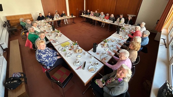 Eine Gruppe von Menschen sitzt an langen Tischen in einer Speisehalle, unterhält sich und genießt gemeinsam eine Mahlzeit.