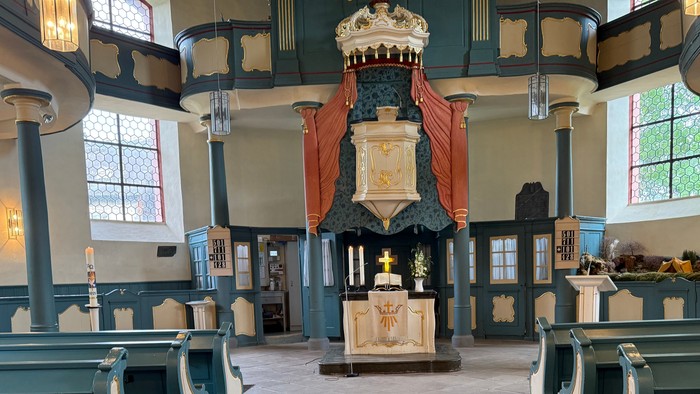 Historisches Kircheninneres mit hölzernen Kirchenbänken, einer verzierten Kanzel und bogenförmigen Buntglasfenstern.