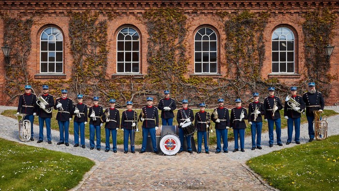 En musikkorps i uniformer spiller foran et rødt murstenshus med bjerke.