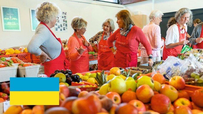 Gruppe von Frauen, die auf einem Markt frische Produkte auswählen, mit einer ukrainischen Flagge im Vordergrund.