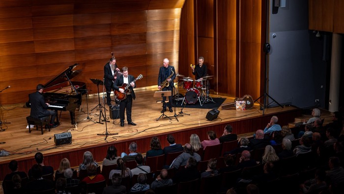 Eine Live-Jazzband tritt auf der Bühne in einem Konzertsaal vor einem aufmerksamen Publikum auf.