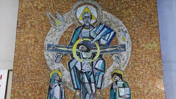 Ein Steinaltar mit Blumenschmuck und zwei Kerzen steht vor einem großen Mosaik-Wandbild. Das Mosaik zeigt zentral die Heilige Dreifaltigkeit.