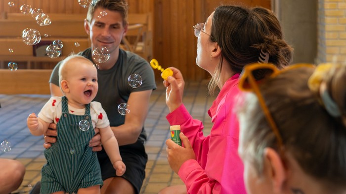 En glad gruppe leger med bobler og legetøj, herunder en baby, der griner omgivet af voksne og børn.