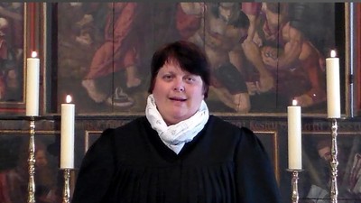 Eine Person in geistlichen Gewändern mit der Aufschrift „Ms Clotilde“, die an einem religiösen Podium mit brennenden Kerzen und Kunstwerken im Hintergrund spricht.