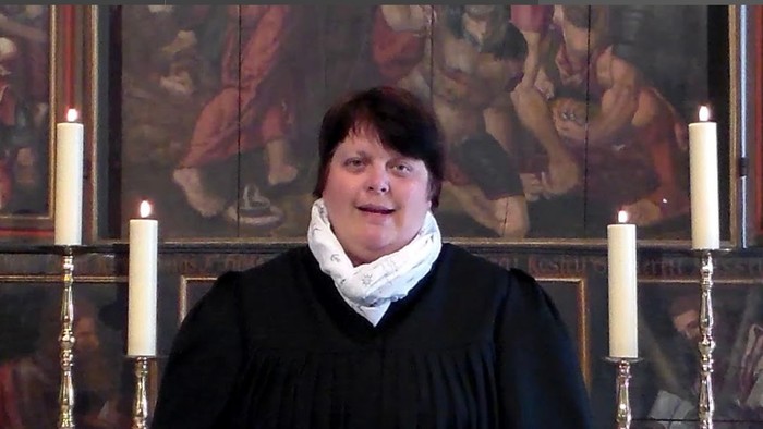 Eine Person in geistlichen Gewändern mit der Aufschrift „Ms Clotilde“, die an einem religiösen Podium mit brennenden Kerzen und Kunstwerken im Hintergrund spricht.