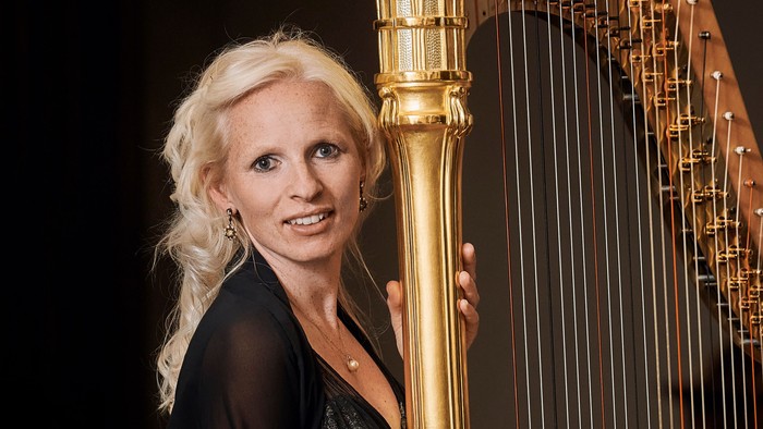 Eine Frau in eleganter Kleidung posiert neben einer großen, verzierten goldenen Harfe.