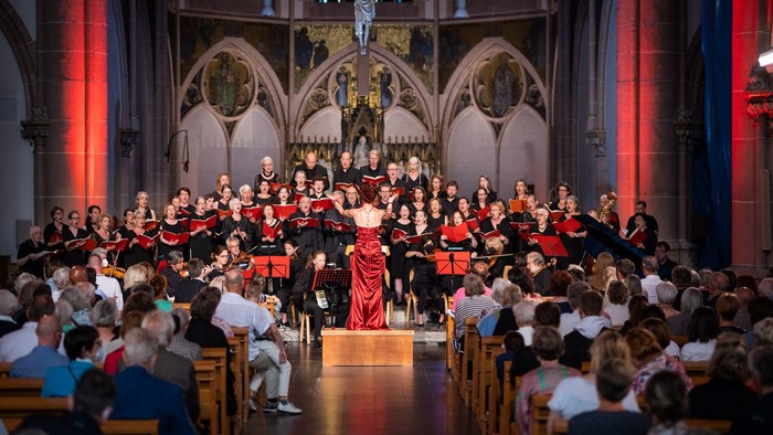 Ein großer Chor tritt in einer prächtigen Kathedrale auf, während ein aufmerksames Publikum in hölzernen Kirchenbänken sitzt.