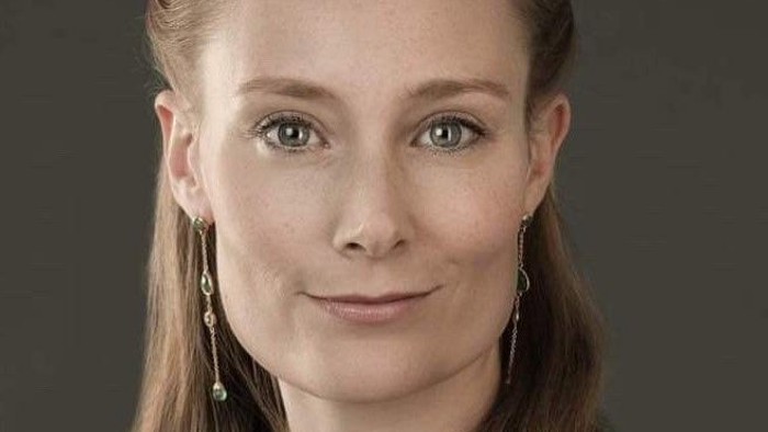 Portræt af en person med langt, glat hår iført dinglende øreringe på en ensfarvet mørk baggrund.