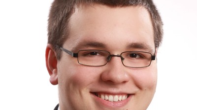 Ein lächelnder Mann in formeller Kleidung mit Brille, der einen Anzug und eine Krawatte trägt.