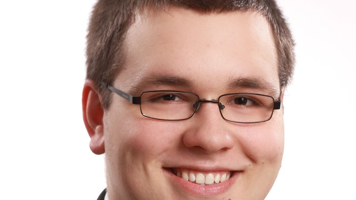 Ein lächelnder Mann in formeller Kleidung mit Brille, der einen Anzug und eine Krawatte trägt.