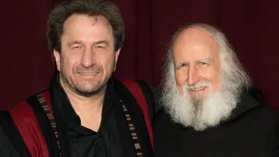 Zwei lächelnde Männer mit Bärten posieren gemeinsam vor einem weinroten Hintergrund.