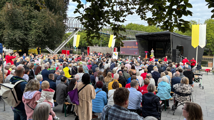 Eine große Menschenmenge versammelt sich im Freien zu einer öffentlichen Veranstaltung oder einem Konzert in einer parkähnlichen Umgebung mit einer Bühne.