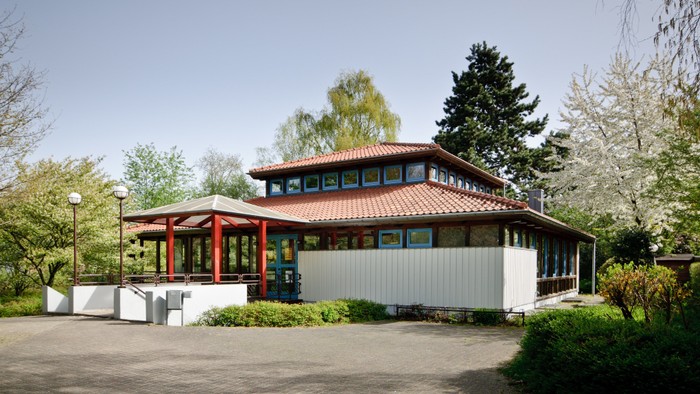 Das Bild zeigt ein modernes Gebäude mit rotem Dach und großen Fenstern, umgeben von Grünflächen und Bäumen.