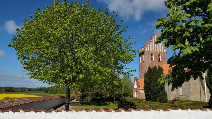En historisk murstens tårn indrammet af frodige grønne træer og en hvid mur med teglstenstag i baggrunden.