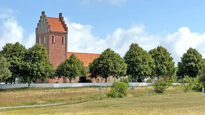 Historisk murstenskirke med højt tårn omgivet af frodige grønne træer og åbne græsarealer.
