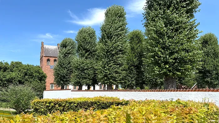 En historisk murstenskirke omgivet af frodig grønne områder og farverigt efterårsløv under en klar himmel.