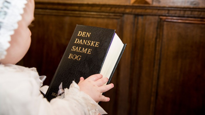 Et spædbarn i hvidt tøj holder en dansk salmebog med titlen *