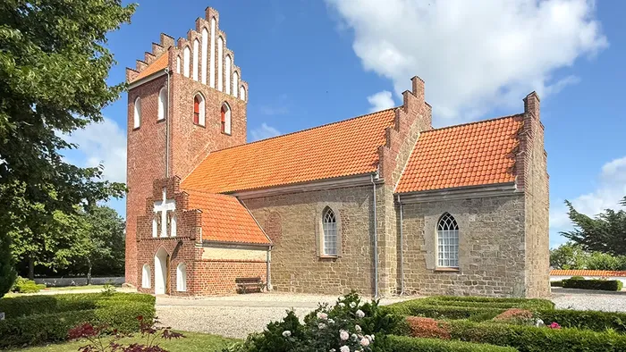 Historisk murstenskirke med rødt teglstenstag og frodige grønne omgivelser, prydet med buede vinduer og et højt tårn.