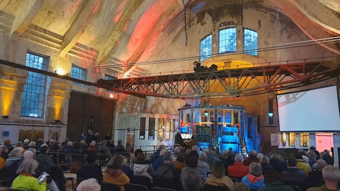 Großer Saal mit Publikum bei einer Veranstaltung, Bühne mit Projektionen und Redner.