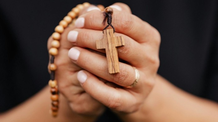 Hände gefaltet, die einen hölzernen Rosenkranz mit Kreuzanhänger in betender Haltung halten.