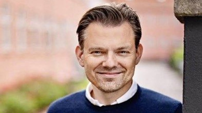 En smilende mand i en mørkeblå sweater, der står udendørs ved siden af en stensøjle.