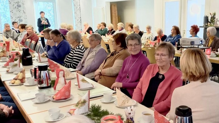 Eine Gruppe von Menschen sitzt in einem gut beleuchteten Raum an Tischen, unterhält sich und genießt eine Mahlzeit.