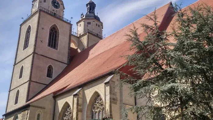 Historische Kirche mit hohem Turm und rotem Ziegeldach, eingerahmt von einem immergrünen Baum und angrenzenden Gebäuden.