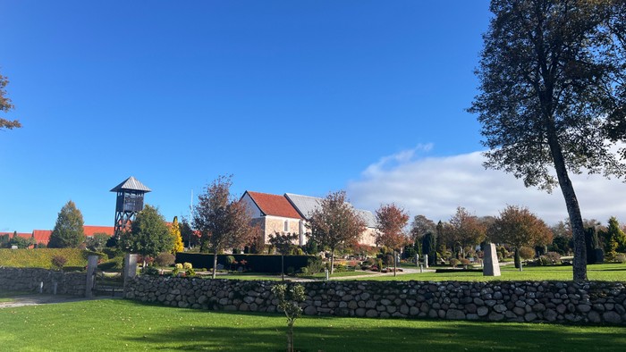 En fredfyldt park med en stenmur, træer og en bygning med rødt tag under en klar, blå himmel.
