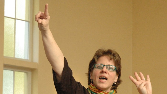 Eine Person mit Brille und Schal gestikuliert begeistert, während sie vor einem Fenster spricht.