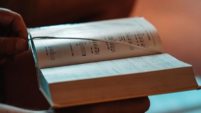 Eine Hand hält ein offenes Buch mit sichtbarer Notenblattmusik, umgeben von anderen Büchern auf einer gemütlichen Oberfläche.