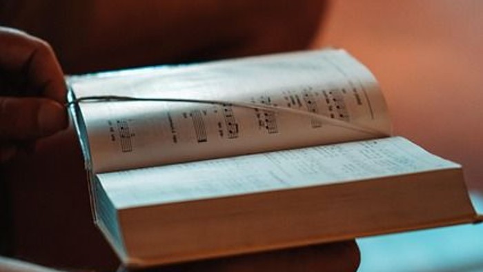 Eine Hand hält ein offenes Buch mit sichtbarer Notenblattmusik, umgeben von anderen Büchern auf einer gemütlichen Oberfläche.