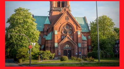 Das Bild zeigt eine rote Kirche mit grünen Dächern und einem Uhrturm, umgeben von Bäumen und Gras.