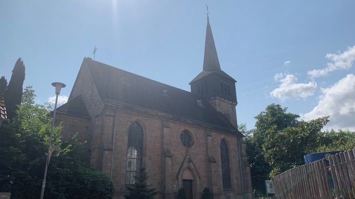 Kirche mit hohem Turmspitz und Rundbogenfenstern, umgeben von Bäumen und klarem Himmel.