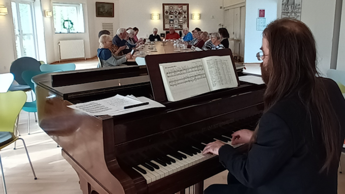 En kvinde spiller piano i en rum med andre musikere og lyttere.