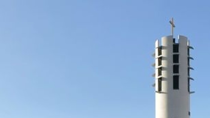 Modernes Gebäude mit einem hohen, zylindrischen Turm, der von einem goldenen Kreuz gekrönt wird, vor klarem Himmel.