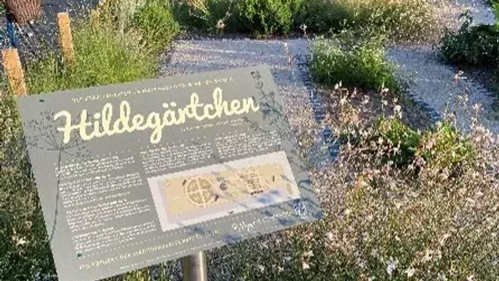 Informationsschild über den Hildegärtchen-Garten in einer gestalteten Außenanlage mit Pflanzen und Wegen.
