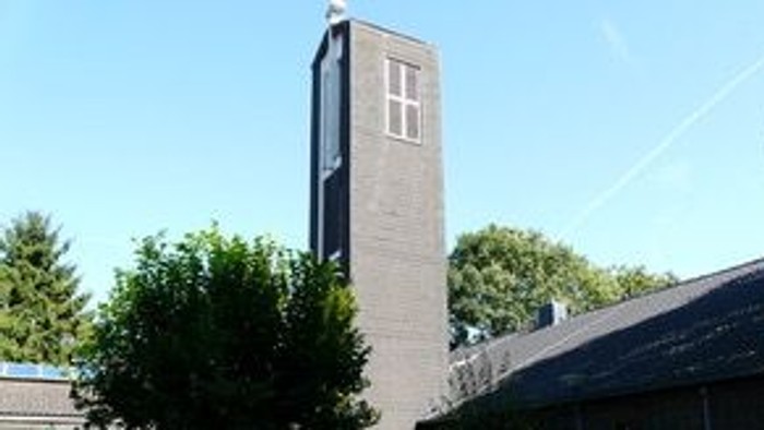 Ein hoher Backsteinkirchturm steht neben einem Gebäude mit grünen Bäumen und Bänken in einem sonnigen Innenhof.