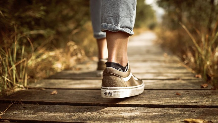 En der går på træbrædder med græs og træer ved siden af. Blå stumpe-jeans anke strømper og brun kondi sko