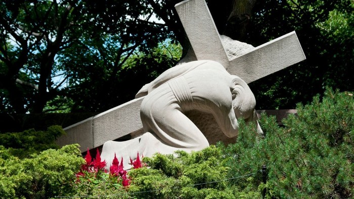Eine feierliche Statue von Jesus am Kreuz, umgeben von üppigem Grün und roten Blumen.